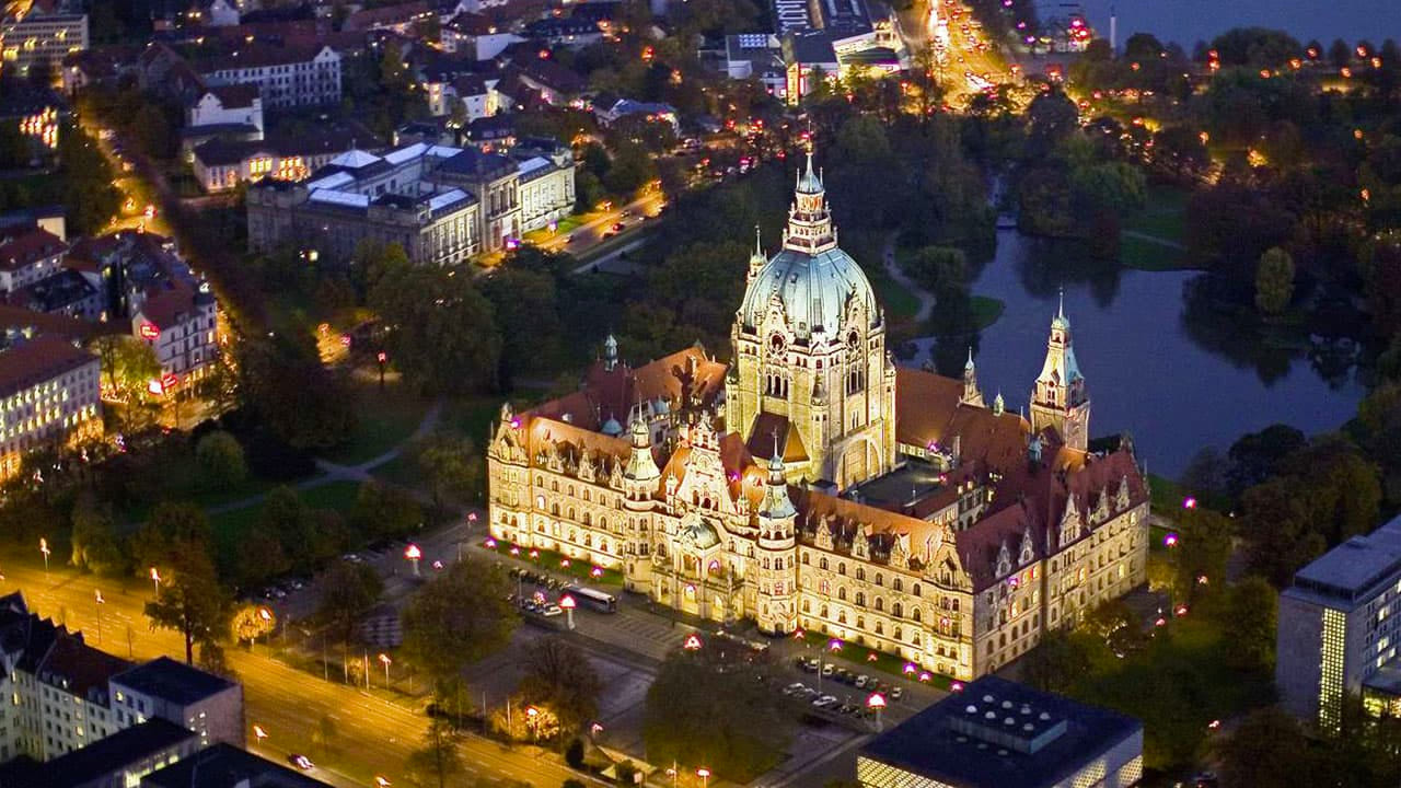 Illuminated New Town Hall in Hanover at night, surrounded by streets, trees, and a park with a lake in the background.