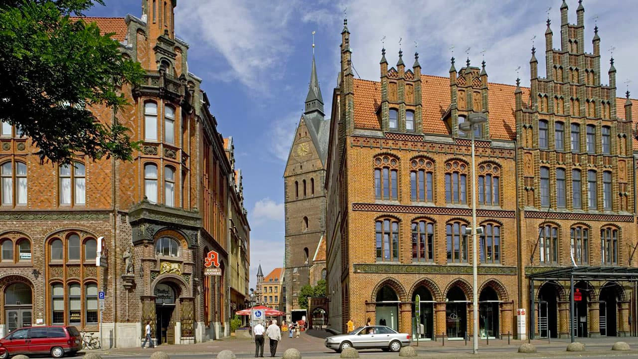 Historic brick façades in Hanover's old town during the day. In the center, the tower of the Marktkirche rises, flanked by two richly decorated buildings in the style of North German Brick Gothic.