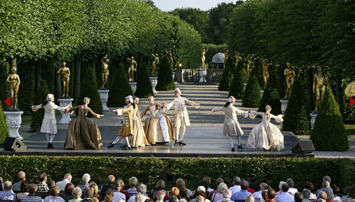 Baroque dance performance in the Great Garden of Herrenhausen in front of an audience, with golden statues and trimmed trees in the background.