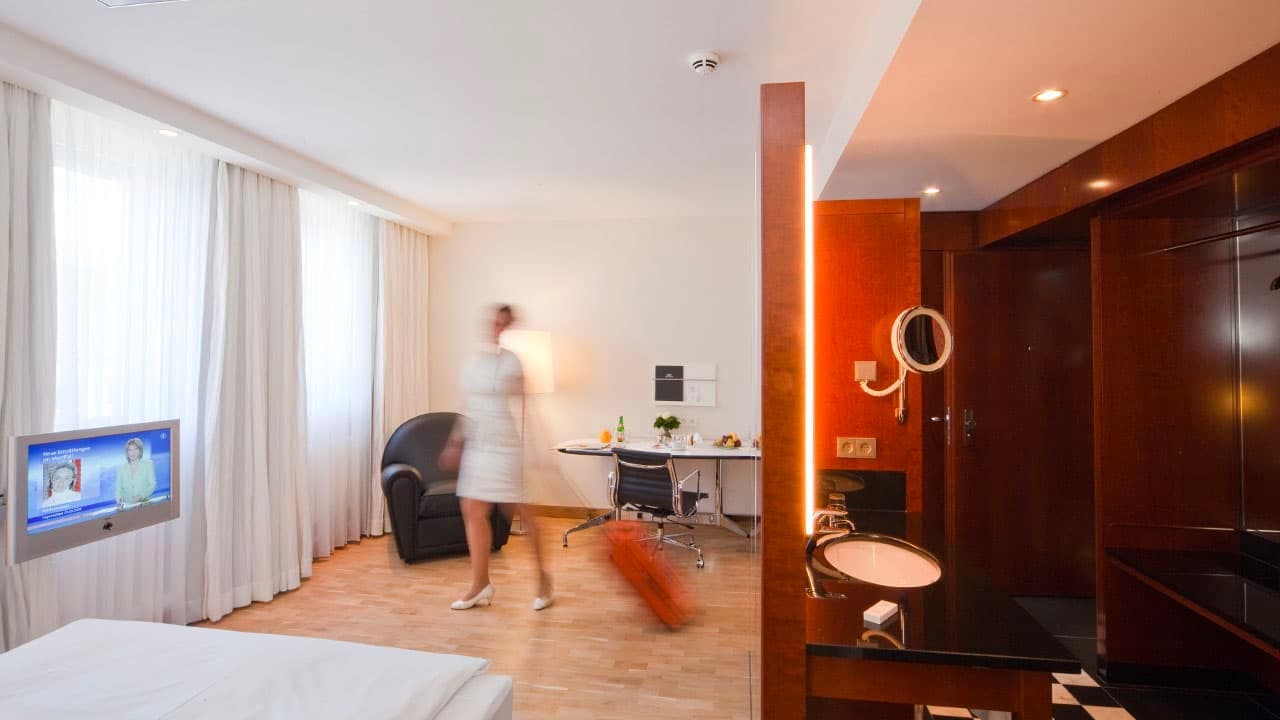 image-room-detail0 Modern single room with bed, TV, desk, and armchair; in the foreground a washstand, in the background a woman with a suitcase in motion.