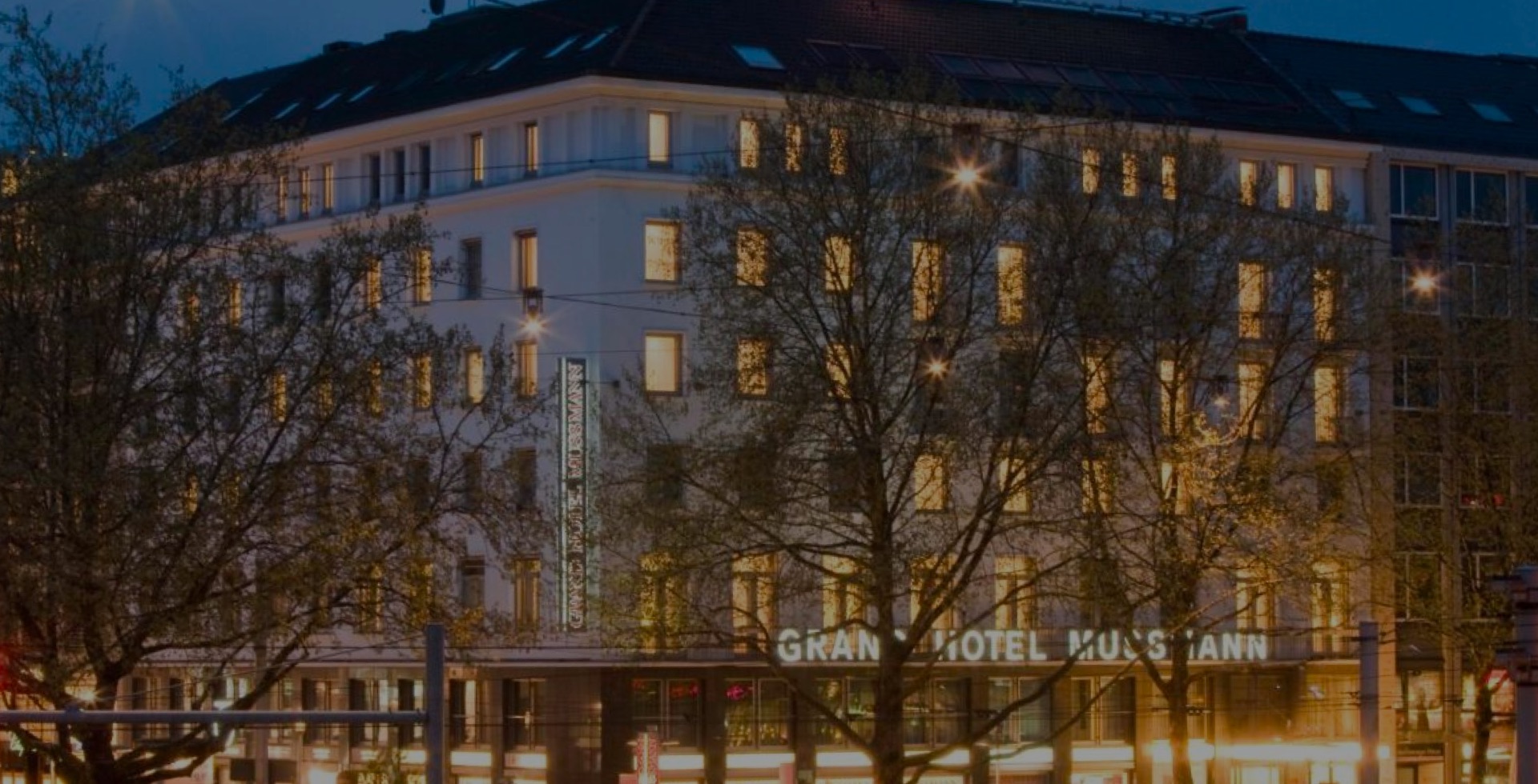 aufmacher-01 Exterior view of the Grand Hotel Mussmann in the evening, illuminated windows and hotel sign, trees in the foreground, dark sky with street lighting.