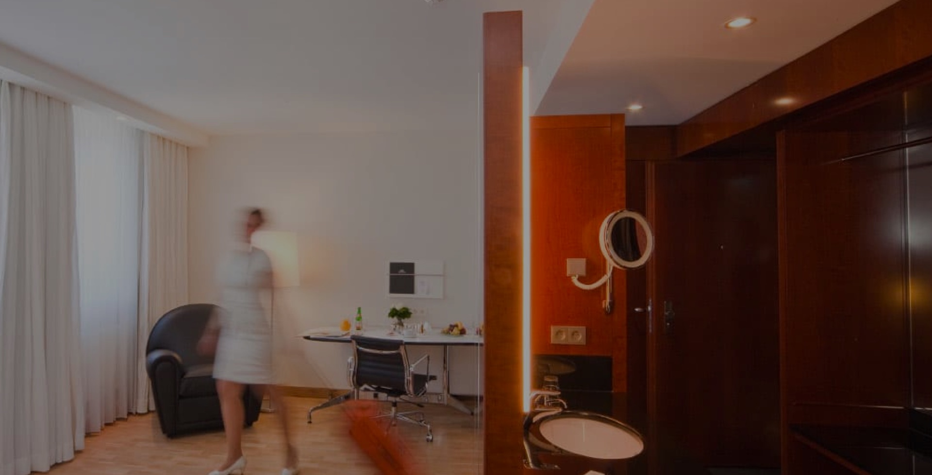 aufmacher-02 Modern hotel room with light curtains, desk and armchair; in the foreground a blurred person in motion, mirror and wash area with dark wood paneling visible.