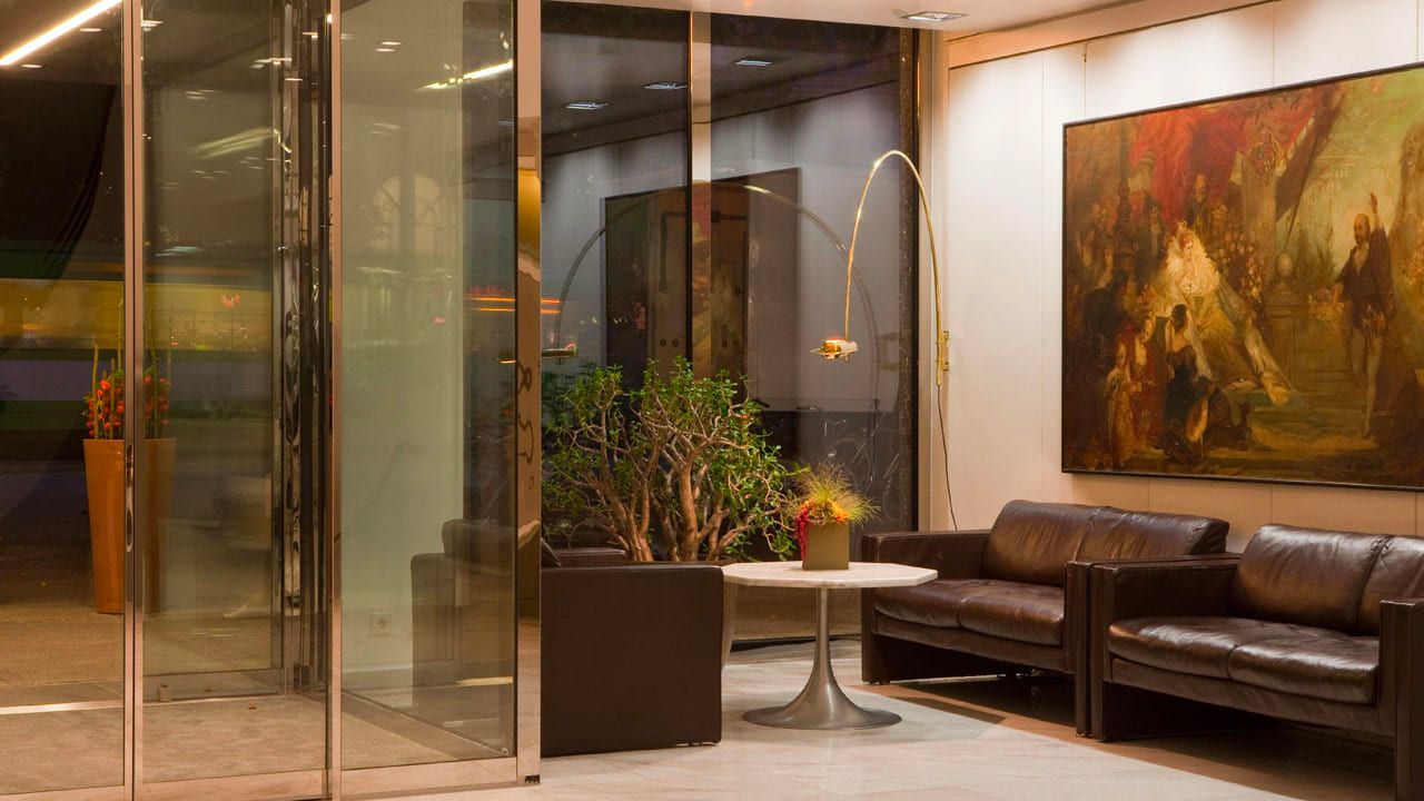 Abendlich beleuchtete Hotellobby mit gläsernem Eingang, Sitzecke mit Ledersofas, rundem Tisch und Dekopflanzen, große Wandmalerei als Blickfang an der Wand.