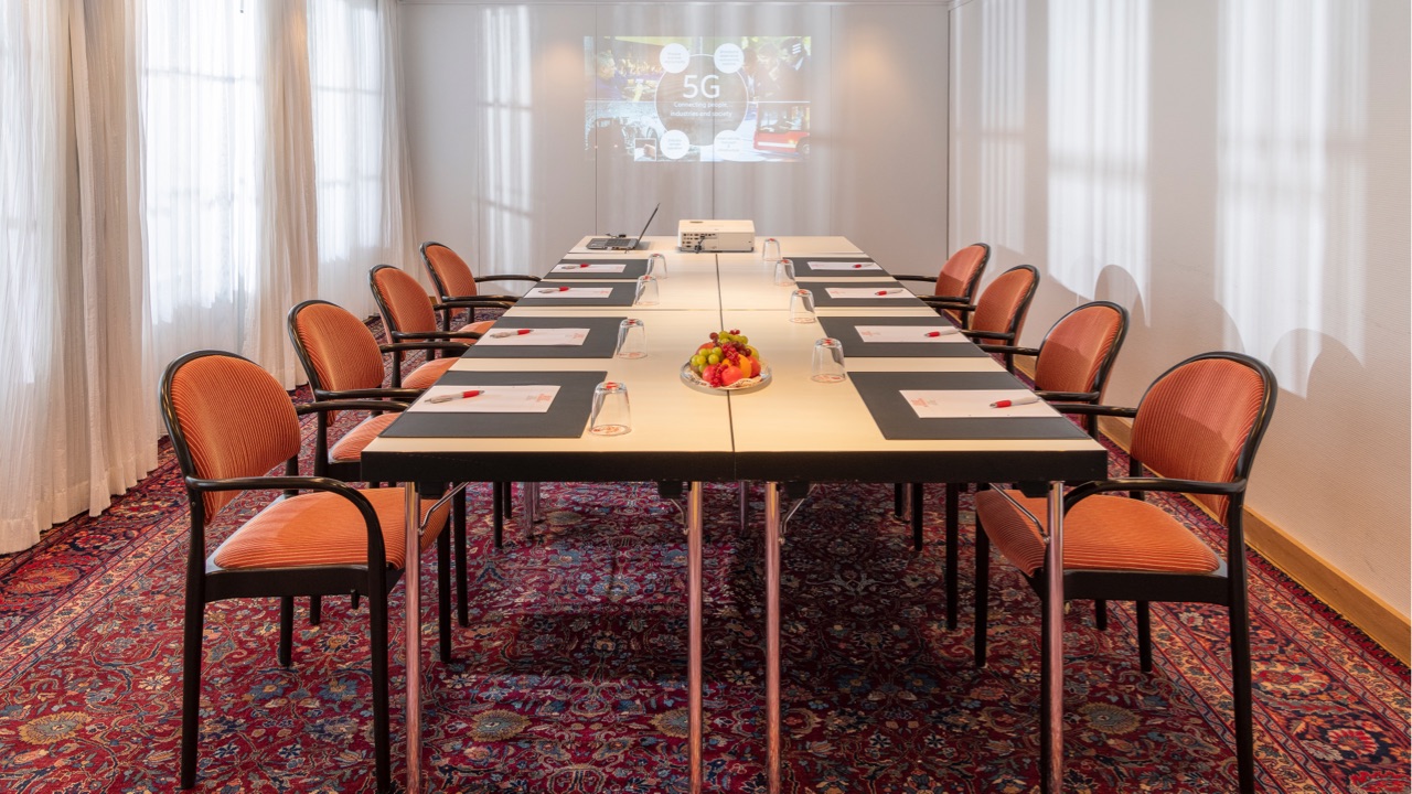 Heller Konferenzraum mit U-förmig angeordneten Tischen, orange gepolsterte Stühle, auf dem Teppichboden stehend, Projektor mit Präsentation an der Wand, Tageslicht durch große Fenster mit Vorhängen.