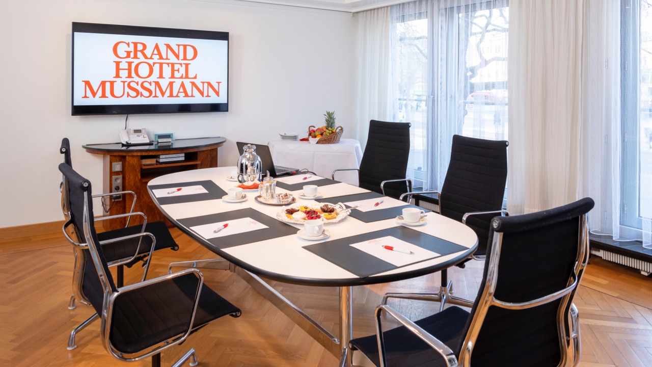 Kleiner Besprechungsraum mit ovalem Tisch und fünf schwarzen Konferenzstühlen, TV-Bildschirm mit Schriftzug 'Grand Hotel Mussmann', gedeckter Tisch mit Kaffee, Tee und Gebäck, Tageslicht durch bodentiefe Fenster.