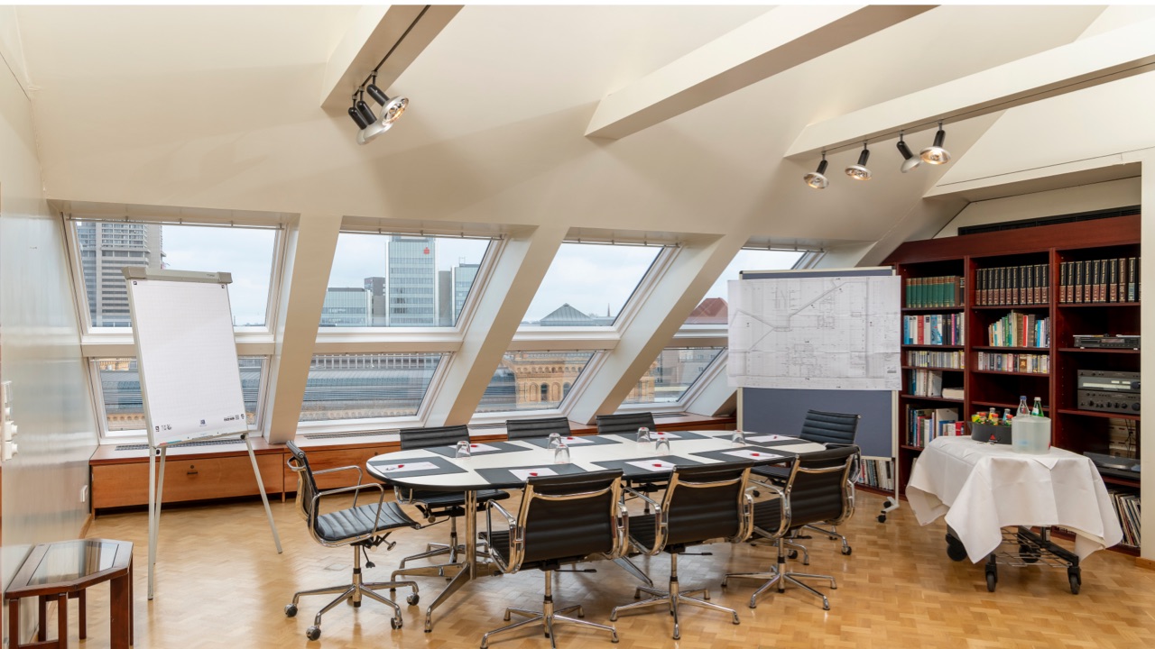 Heller Konferenzraum unter dem Dach mit großer Fensterfront, Blick auf die Stadt, moderner Besprechungstisch mit acht schwarzen Drehstühlen, Flipchart, Bücherregal und Getränketisch.