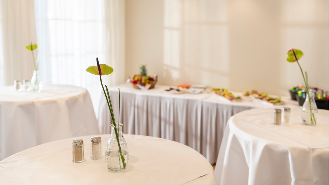 Lichtdurchfluteter Raum mit weiß eingedeckten Stehtischen, einzelne Blumenvasen mit Anthurien als Tischdeko, im Hintergrund ein Buffet mit Snacks, Obst und Getränken.