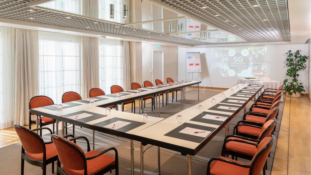 Geräumiger Konferenzraum mit Tischen in U-Form, rote Polsterstühle, Projektor-Präsentation zum Thema 5G an der Wand, Flipcharts im Hintergrund, große Fensterfront mit lichtdurchlässigen Vorhängen.