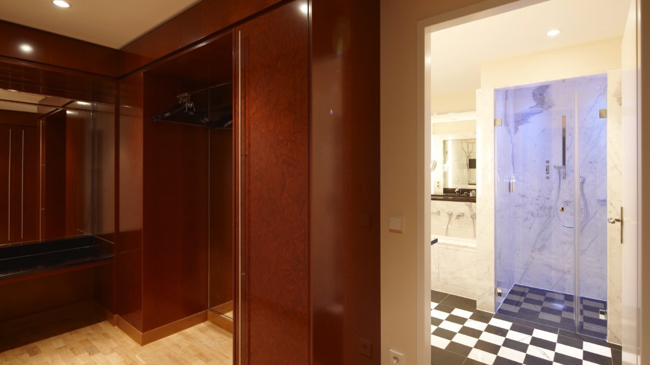 Blick aus einem begehbaren Holzkleiderschrank in ein elegantes Bad mit Marmor-Dusche und schwarz-weißem Fliesenboden.