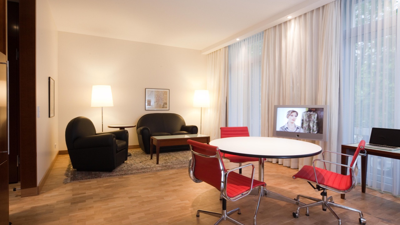 Grand-Hotel-Mussmann-Zimmer-Grand-Suite-02 Wohnbereich einer Hotelsuite mit schwarzem Sofa, Sesseln, rundem Tisch mit roten Stühlen, Fernseher und Schreibtisch am Fenster.