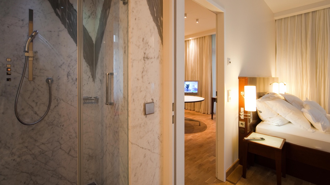 Grand-Hotel-Mussmann-Zimmer-Grand-Suite-04 Blick aus dem Badezimmer mit Marmor-Dusche in ein modernes Hotelzimmer mit Bett, Fernseher und Schreibtisch im Hintergrund.