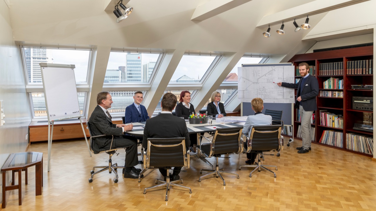 Geschäftstreffen in einem hellen Konferenzraum mit sieben Personen und Stadtblick; ein Mann präsentiert einen Plan.