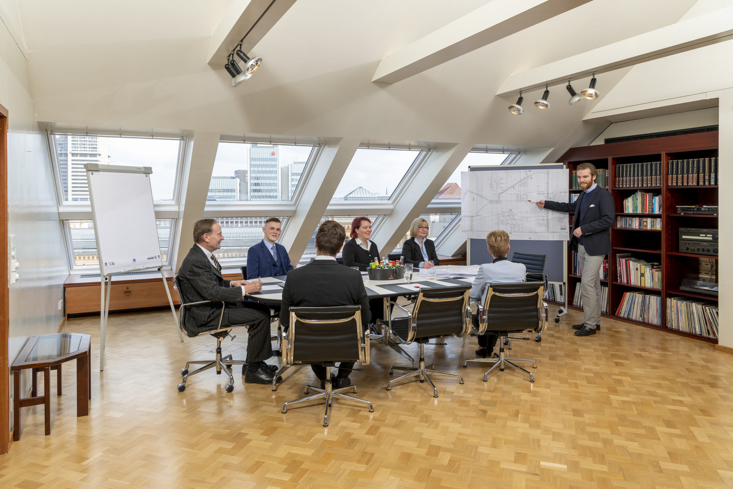 DE: Geschäftstreffen in einem hellen Konferenzraum mit sieben Personen und Stadtblick; ein Mann präsentiert einen Plan. / EN: Business meeting in a bright conference room with seven people and a city view; a man is presenting a plan.