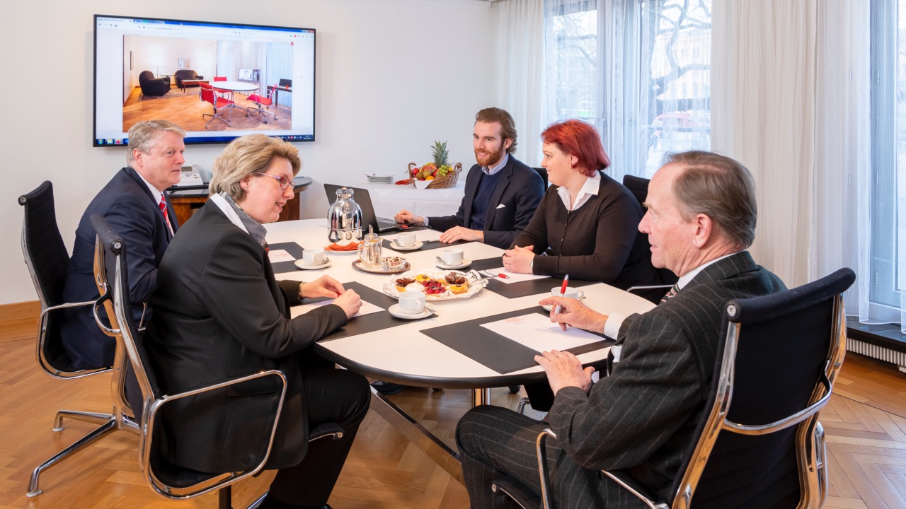 DE: Sechs Personen im Business-Outfit bei einem Meeting an einem Konferenztisch mit Kaffee und Gebäck. / EN: Six people in business attire at a meeting around a conference table with coffee and pastries.