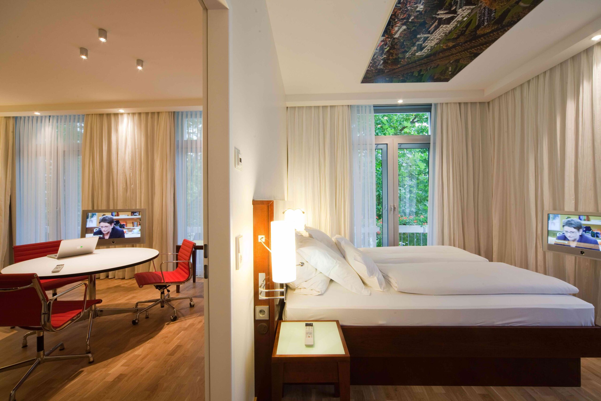 Blick in eine moderne Hotelsuite mit Schlafzimmer und separatem Arbeitsbereich, ausgestattet mit roten Designerstühlen, Schreibtisch und zwei Fernsehern.