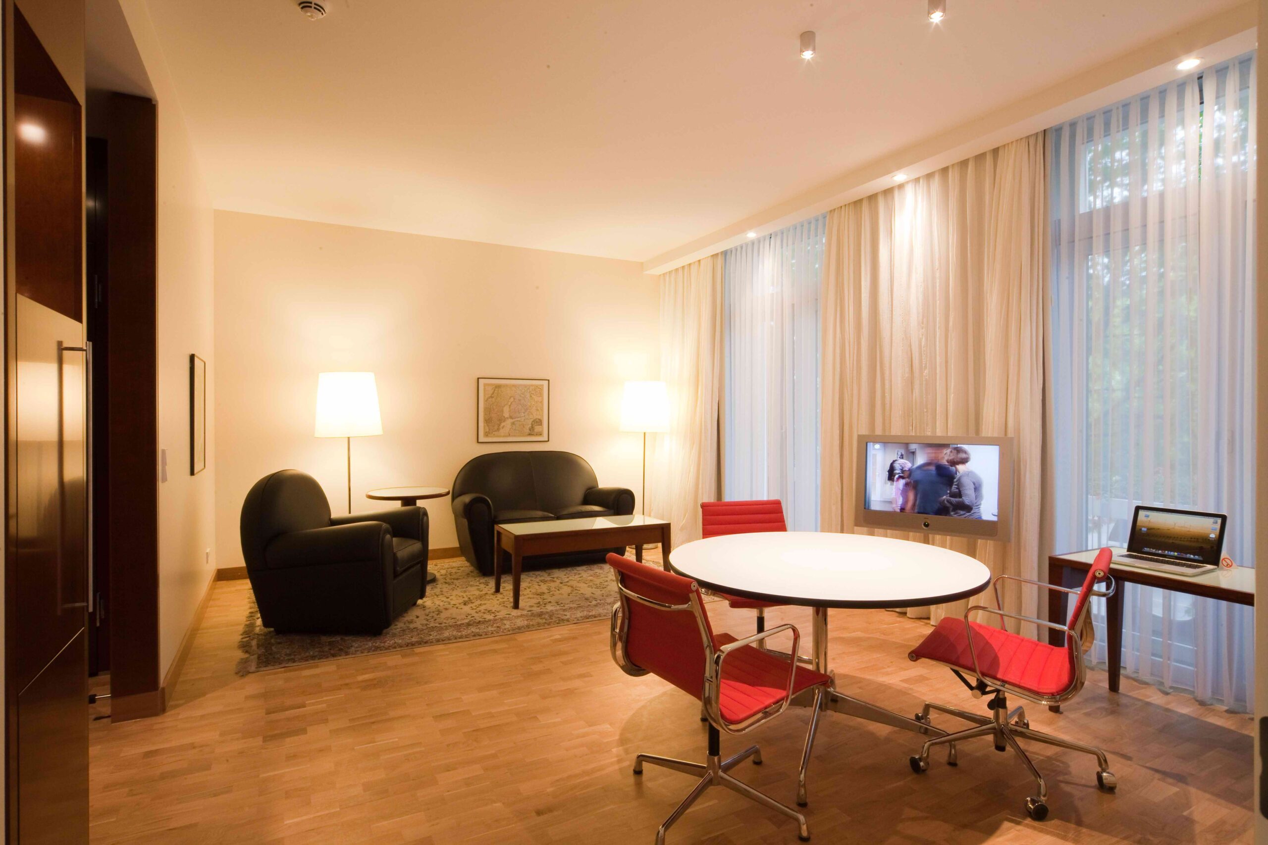 Wohnbereich einer Hotelsuite mit schwarzem Sofa, Sesseln, rundem Tisch mit roten Stühlen, Fernseher und Schreibtisch am Fenster.
